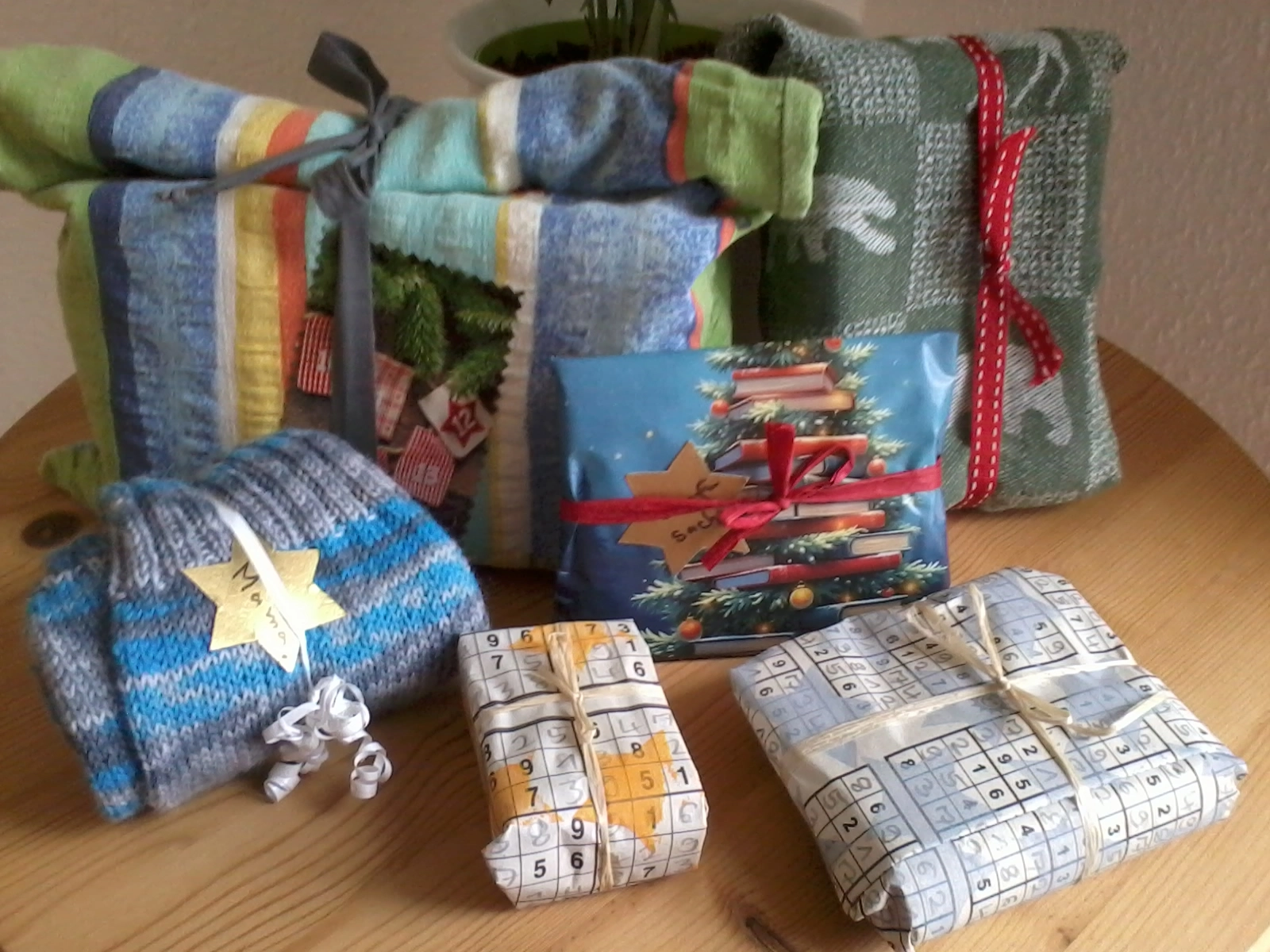 Das Foto zeigt unterschiedlich eingepackte Geschenke: Ein Geschenk ist einem bunten Stoffbeutel eingepackt, ein anderes in einem grünen Tuch mit einer roten Schleife. Zwei Geschenke sind in selbst bedrucktem Papier eingewickelt. Im Vordergrund liegen zusammengerollte Socken, um die ein Geschenkband gebunden ist.