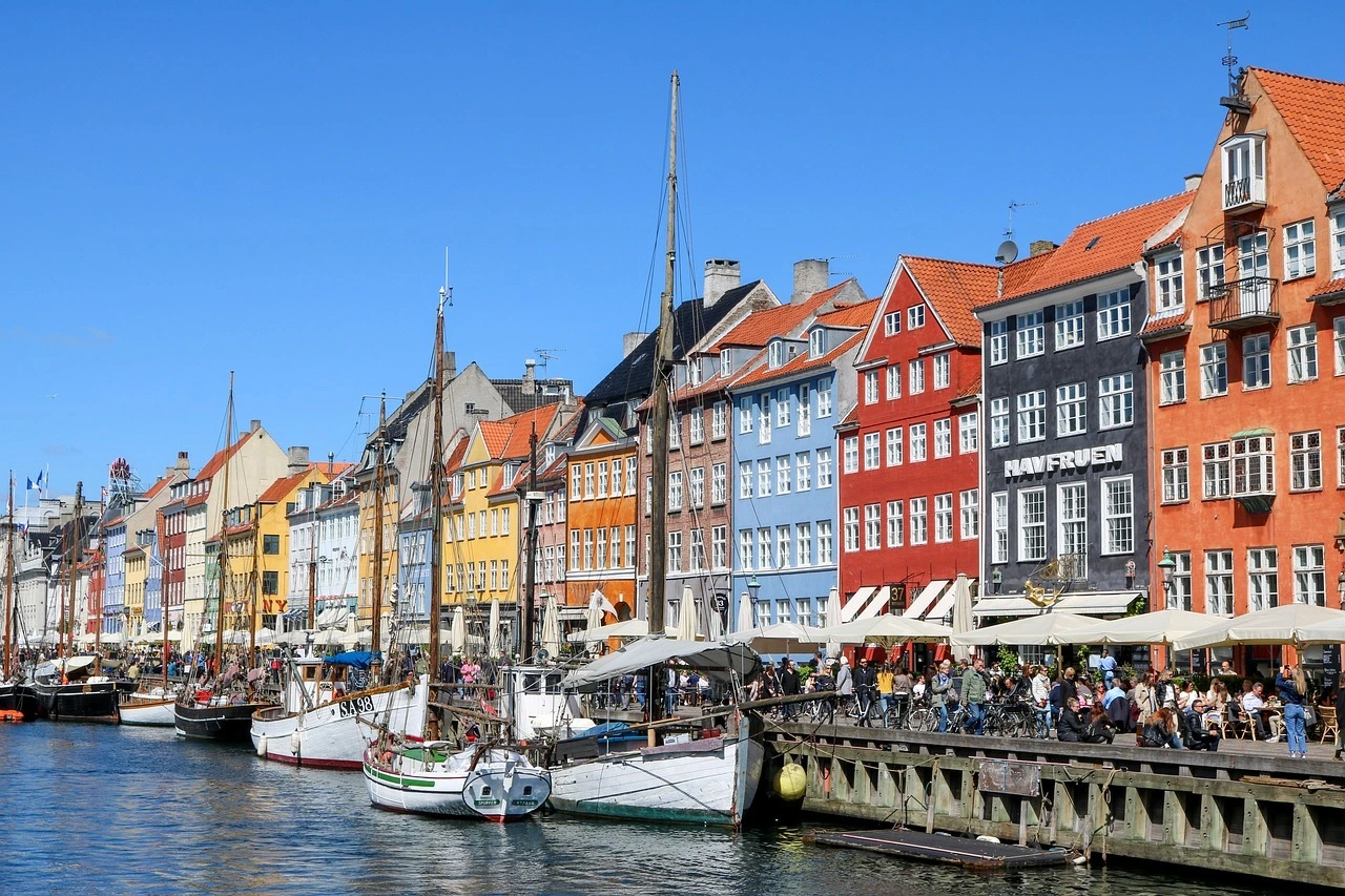 Das Foto zeigt eine Straßenansicht in Kopenhagen. Zu sehen sind mehrere bunte Häuser . Im Vordergrund ist Wasser, darauf Segelboote.