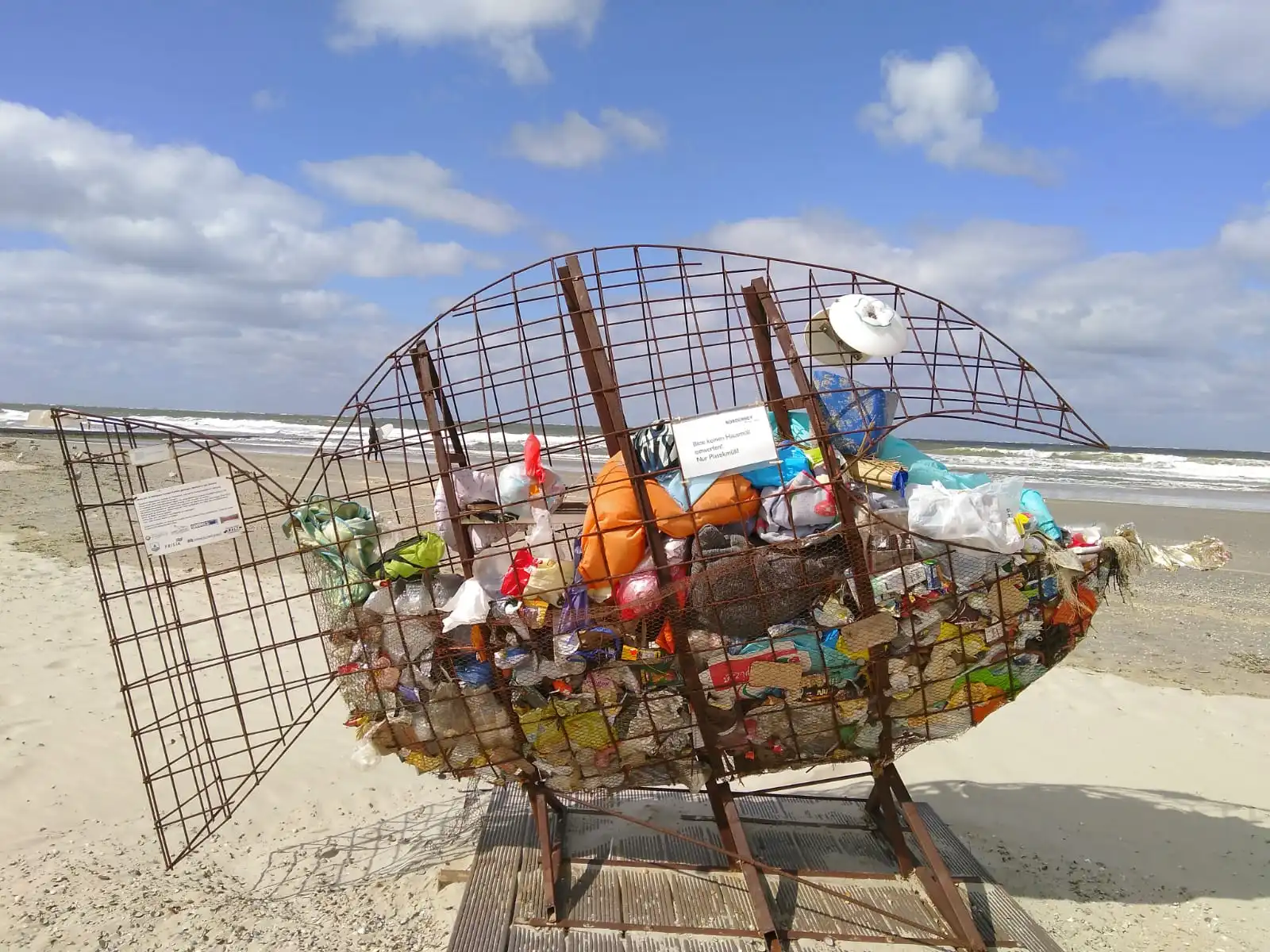 Das Bild zeigt einen Mülleimer am Strand in Form eines riesigen Fischs, der mit Müll gefüllt ist.