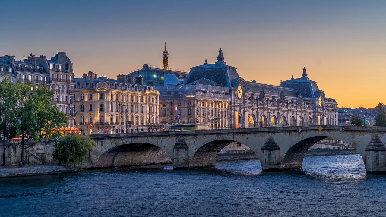 Das Foto zeigt eine Stadtansicht von Paris im Abendlicht. Im Vordergrund ist ein Fluss mit einer Brücke zu sehen.