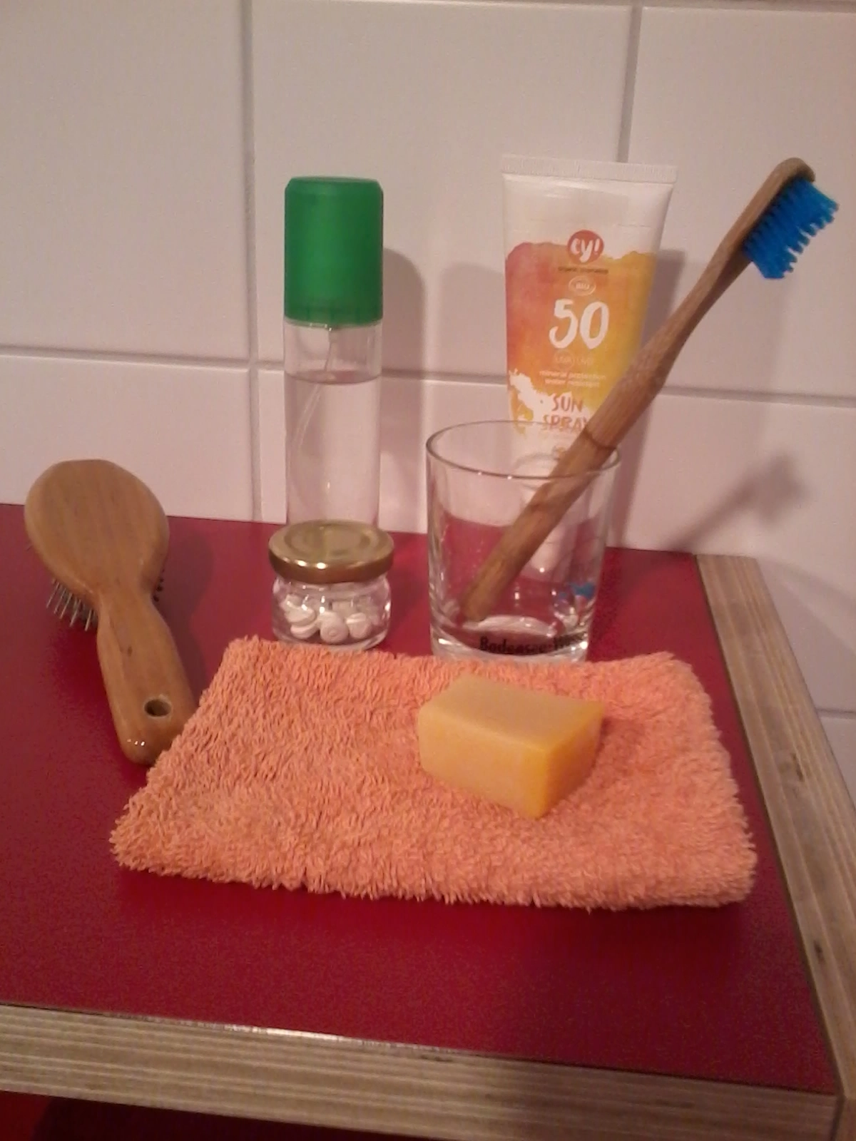 Das Foto zeigt eine feste Haarseife, ein kleines Gläschen mit Denttabs, eine Bmabus-Zahnbürste in einem Zahnputzglas, eine Haarbürste aus Holz und eine Deo-Flasche.
