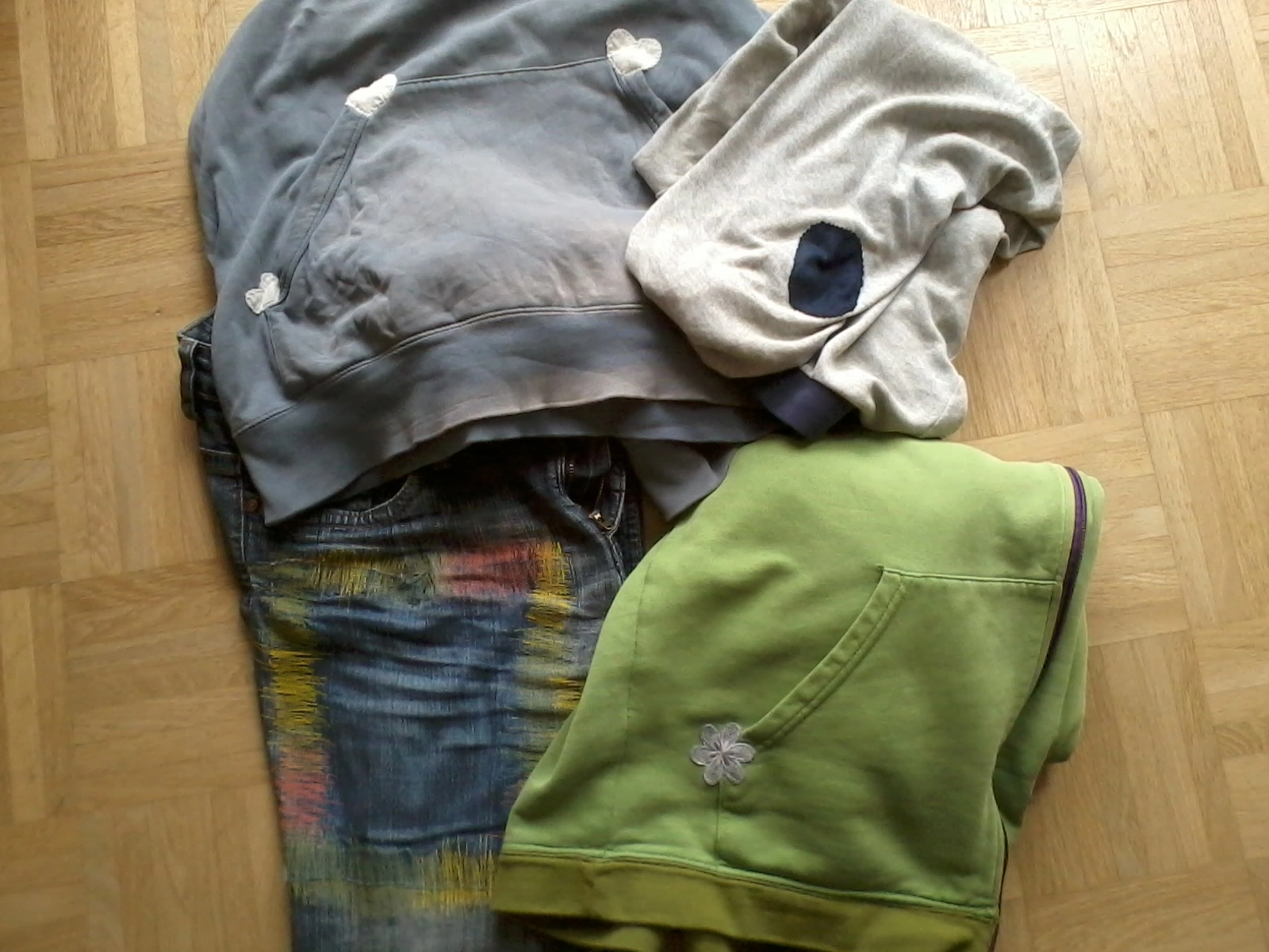 Vier Kleidungsstücke mit Flicken: Ein hellgraues Oberteil, eine Jeans, eine hellgrüne Jacke und ein graues Shirt.