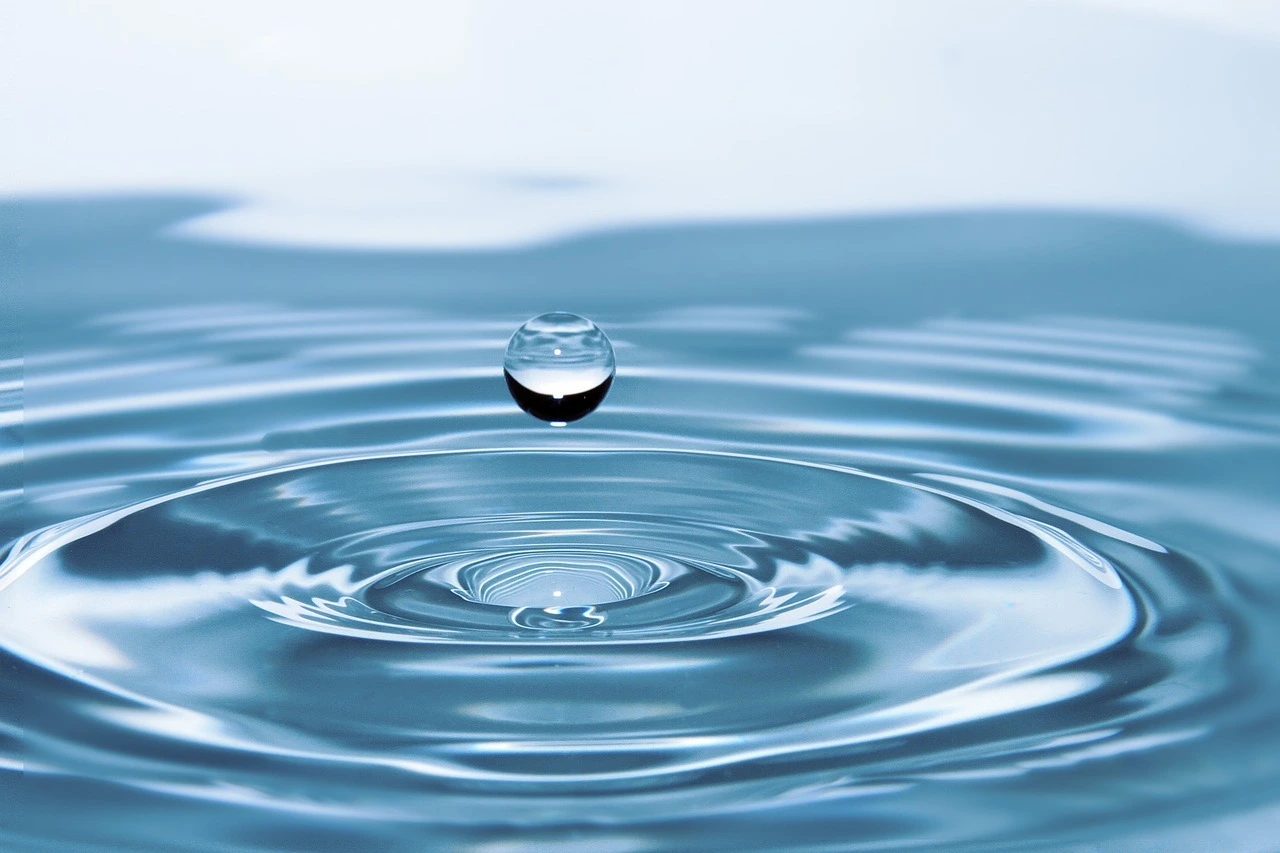 Das Foto zeigt eine Wasserfläche. Ein Tropfen Wasser fällt auf das Wasser.