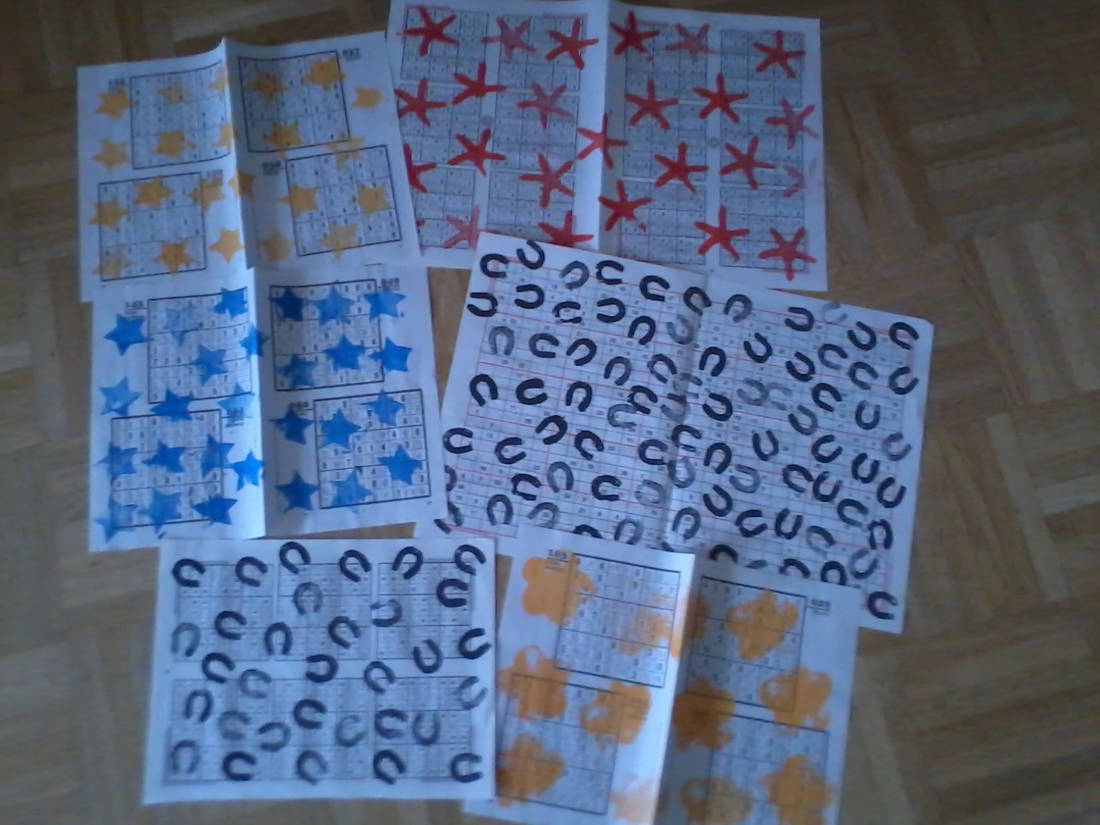 Das Foto zeigt mehrere Seiten aus einem Sudoku-Heft. Sie sind mit bunten Motiven bedruckt: Sterne, Hufeisen, Blumen und liegen auf einem Holzboden.