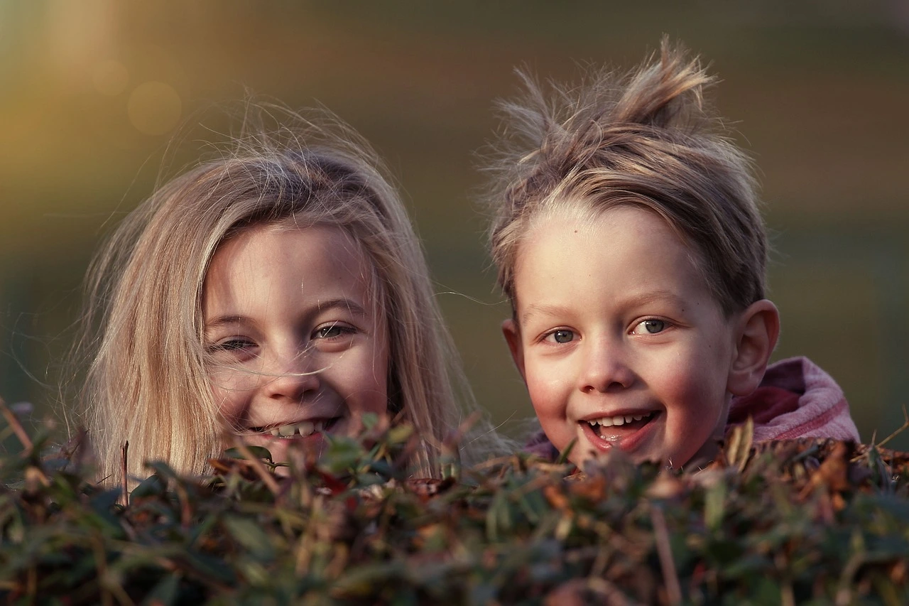 Das Foto zeigt zwei Kinder. Sie schauen hinter einer Hecke vor. Es sind nur die Köpfe zu sehen.
