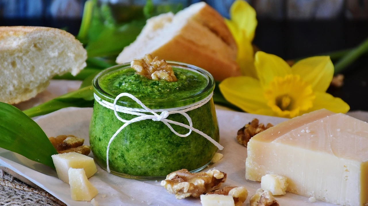 Das Foto zeigt ein Glas mit grünem Pesto sowie ein Stück Brot, Käse und einige Nüsse.