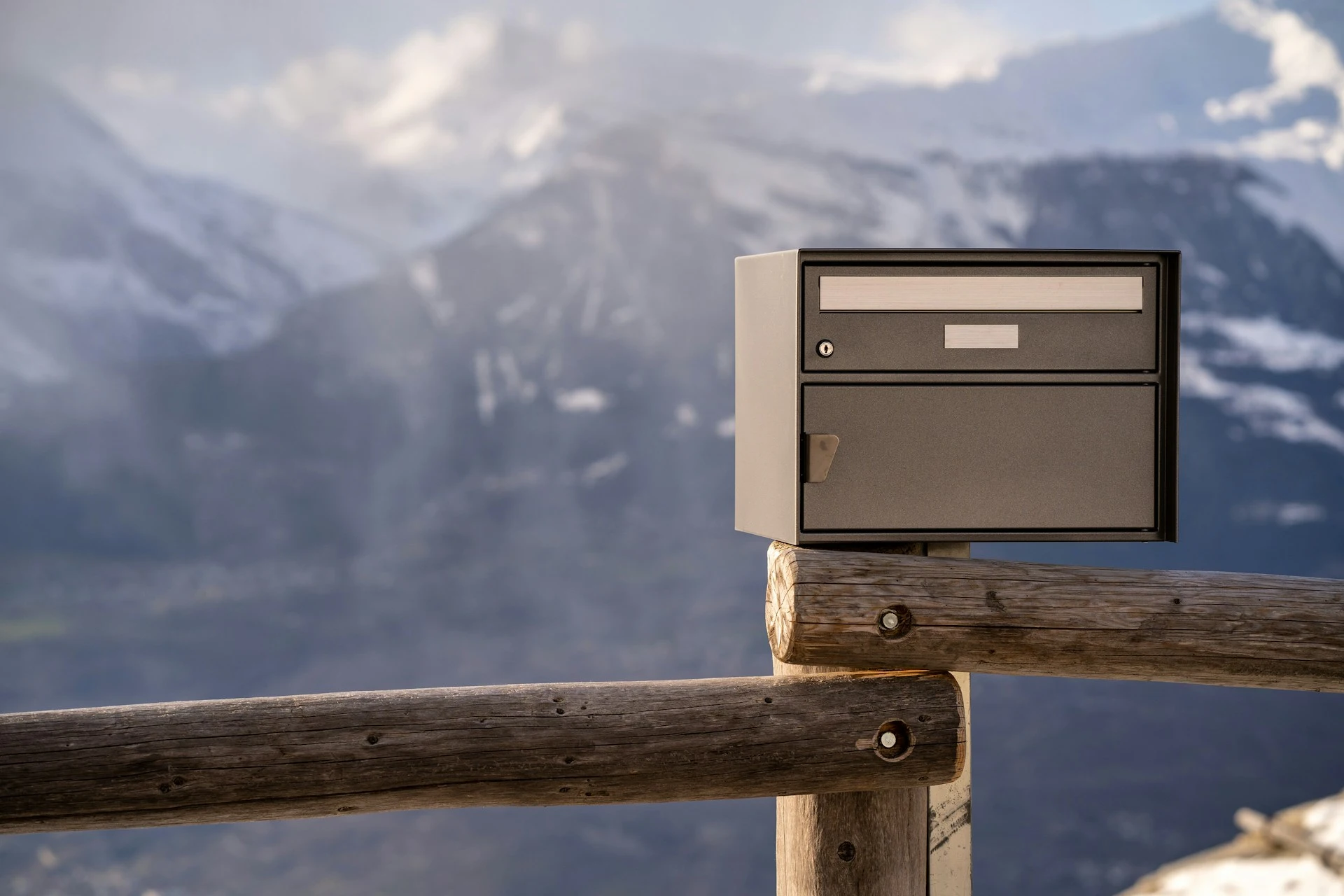 Ein grauer Briefkasten auf einem Holzbalken, im Hintergrund ein Bergmassiv