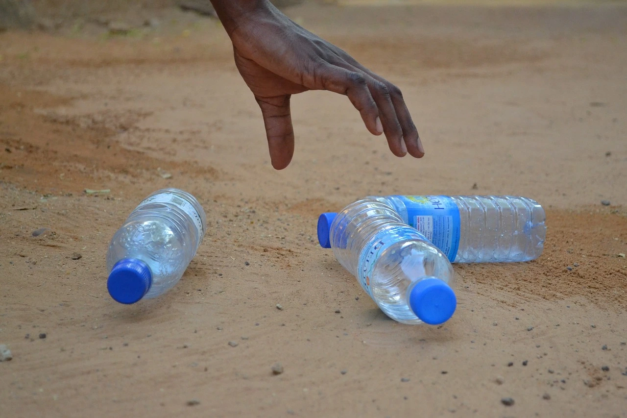 Das Foto zeigt einen Sandstrand mit 3 Plastikflaschen. Eine Hand greift nach den Flaschen.