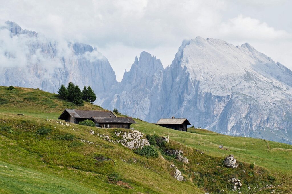 Drei Almgebäude liegen geduckt zwischen grasbewachsenen Hügeln, im Hintergrund felsiges Hochgebirge