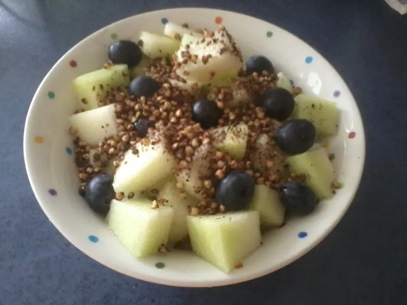 eine Müslischale, gefüllt mit Trauben und geshnittener Melone, darauf Buchweizen-Granola, steht auf einer dunkelblauen Arbeitsplatte