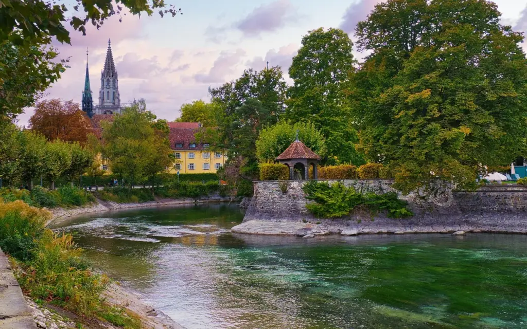 Blick in die Altstadt von Konstanz, im Vordergrund eine Wasserfläche, im Hintergrund das Münster und ein Haus mit gelber Fassade, umgeben von Bäumen