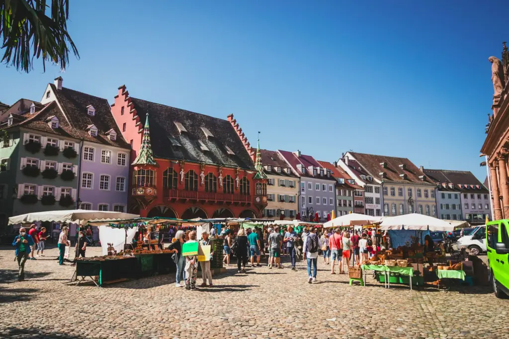Blick auf den Münstermarkt Freiburg, im Vordergrund Menschen und Marktstände, im Hintergrund historische, bunte Gebäude
