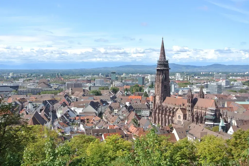 Stadtansicht von Freiburg, im Vordergrund das Münster, umgeben von Hausdächern, im Hintergrund ein MIttelgebirge und Himmel