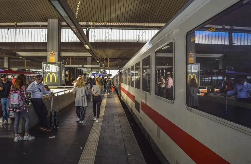Mehrere Menschen stehen und gehen auf einem Bahngleis in einem Bahnhof, rechts steht ein weiß-roter ICE