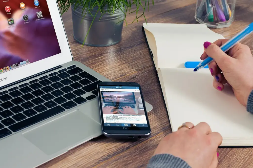 ein Laptop, ein Smartphone sowie ein Notizbuch und ein Glas mit Stiften auf einem Holztisch