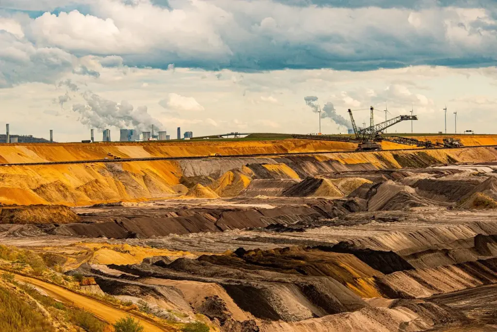 Blick auf einen Kohle-Tagebau, im Hintergrund Maschinen zur Kohleförderung und Kraftwerkstürme, im Vordergrund abgebaute Kohle