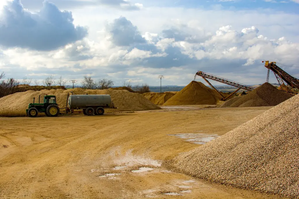 Das Bild zeigt eine Abbaustelle mit mehreren Haufen Kies, Baggern und einem Traktor mit Anhänger.