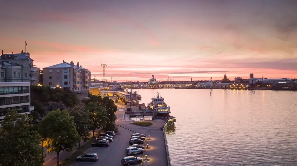 Abendstimmung in einer Stadt an einem Meeresarm, links Gebäude, Bäume und ein Parkplatz, rechts Wasser