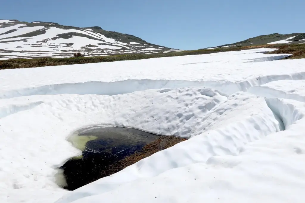 eine Schneefläche, in der Mitte ein dunkler See mit Schmelzwasser, im Hintergrund Berge