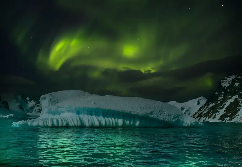 hellgrüne Lichter an einem sehr dunklen Himmel, im Vordergrund ein Eisberg und türkisblaues Wasser