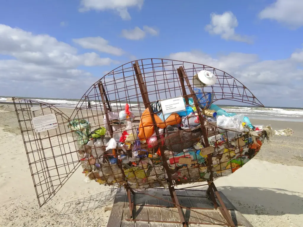 Das Bild zeigt einen großen Mülleimer am Strand in Form eines Fischs, der mit Müll gefüllt ist.