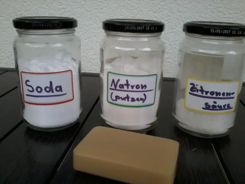 Drei Gläser mit Soda, Natron und Zitronensäure stehen auf einem dunklen Tisch, davor liegt ein Stück Seife