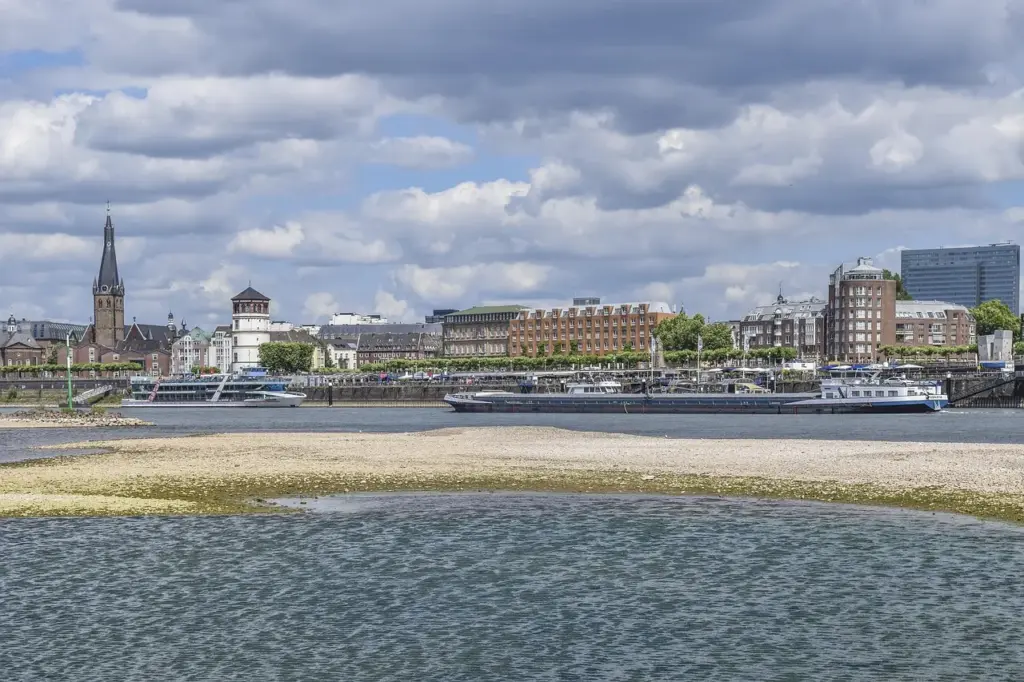 Ein Fluss mit Niedrigwasser, zwischen den wasserführenden Bereichen ist deutlich eine Kiesfläche zu sehen. Im Hintergrund größere Schiffe sowie Häuser einer Stadt