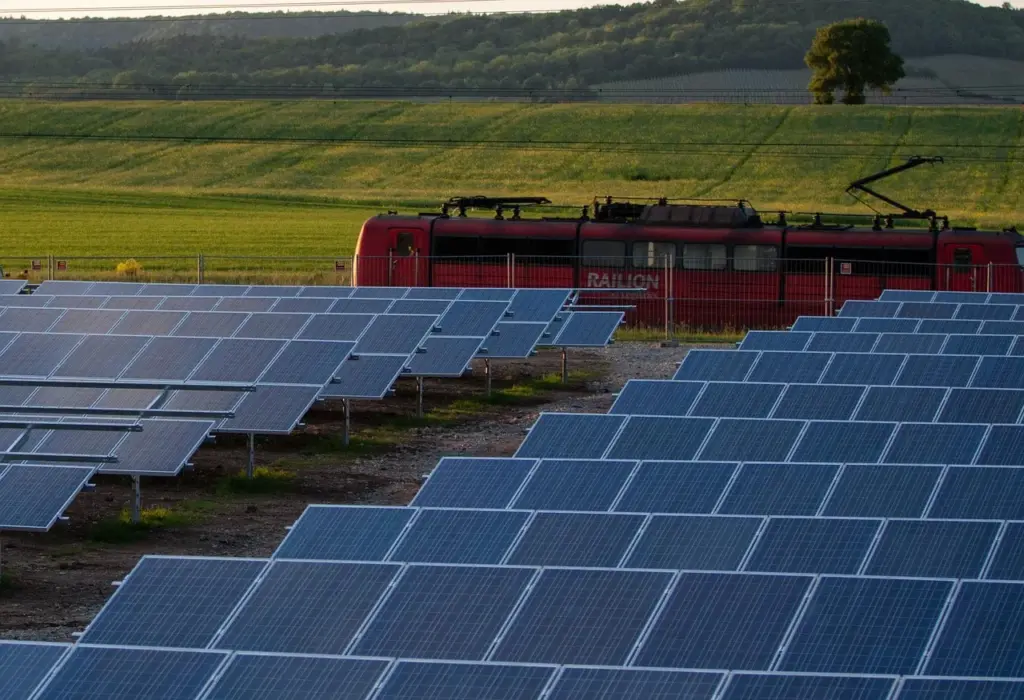 Mehrere Reihen mit Solarzellen stehen auf einer Grünfläche. Im Hintergrund fährt ein Zug vorbei