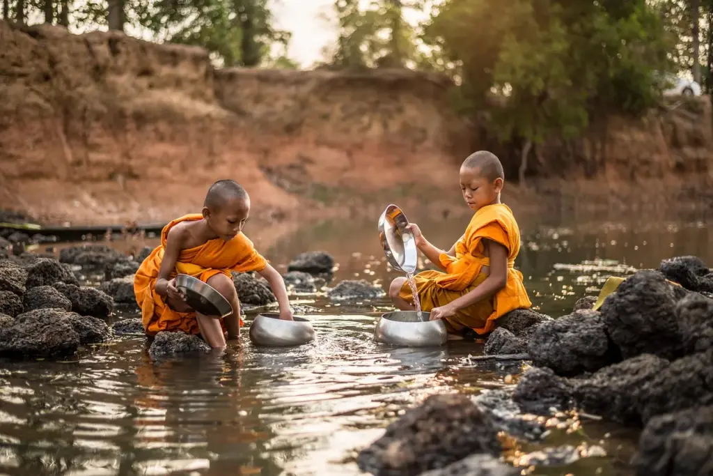 zwei Jungen in buddhistischer Mönchskleidung sitzen auf Steinen in einem Fluss und schöpfen Wasser in Gefäße