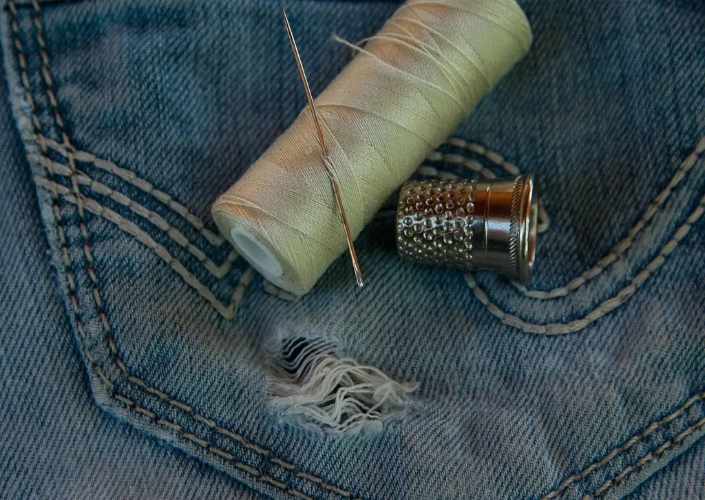 Nahaufnahme einer Jeans mit Loch in der Gesäßtasche, darauf liegen eine Rolle helles Nähgarn, eine Nähnadel und ein Fingerhut aus Metall