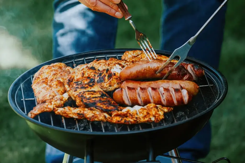 auf einem Grill liegen verschiedene Würstchen und Fleischstücke. Eine Person wendet eine Wurst mit zwei Gabeln